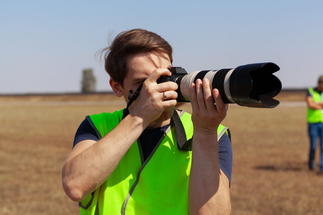 man wearing high visibility vest in fluoro yellow stands outside and points large camera with zoom lens upwards to the sky
